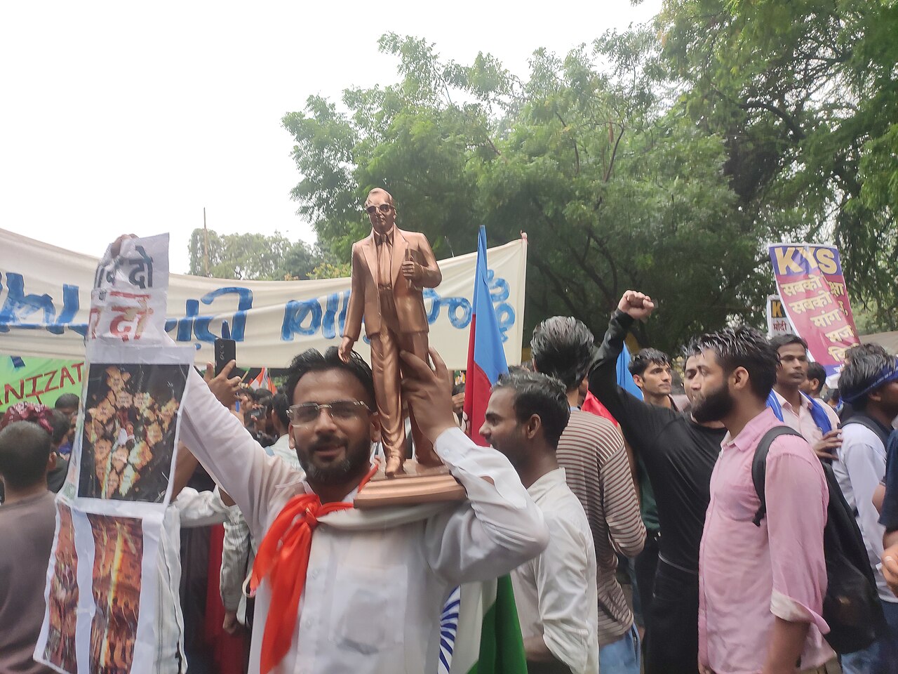 A dense protest crowd at Jantar Mantar; in the foreground a man carries a bronze statue of Dr. B. R. Ambedkar on his shoulder, surrounded by flags, banners and placards