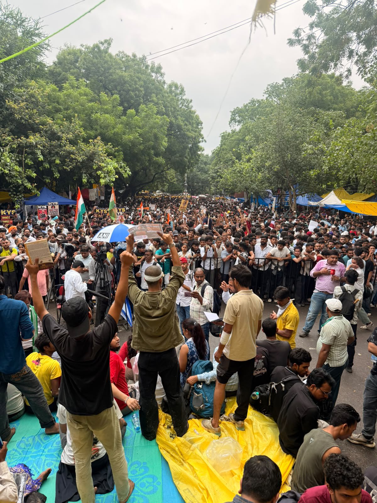 The Jantar Mantar avenue packed with thousands of protesters between rebuilt tarpaulin shelters, tricolour flags flying, protesters in the foreground with arms raised